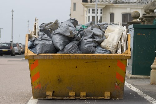 A large yellow industrial skip filled with tightly packed black and white rubbish bags, positioned on a paved street near a row of parked cars and urban buildings in the background. The skip shows signs of dirt and use, with reflective orange and red stripes on its sides. The environment appears to be an outdoor area in a town, possibly near residential or commercial properties, with the rubbish ready for collection. The scene reflects a typical waste disposal setup that Garden Clearance Blackfriars might handle during rubbish removal services in the area, supporting local rubbish clearance needs. The clear weather and natural daylight highlight the details of the bags' textures and the skip's weathered surface, emphasizing the practical aspect of waste management in the Blackfriars district.
