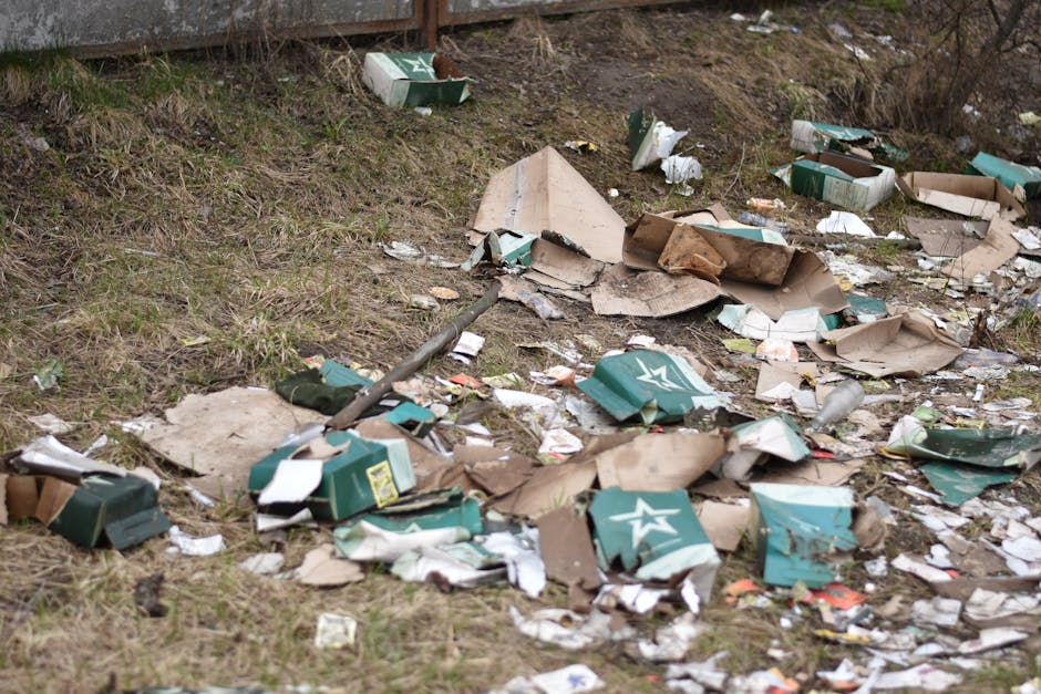 A patch of ground near a grassy slope, scattered with discarded cardboard boxes, some torn and flattened, alongside crumpled paper and plastic packaging. The boxes are mainly green and white, with vis
